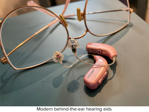 a pair of very modern pink behind-the-ear hearing aids with a pair of glasses in the background