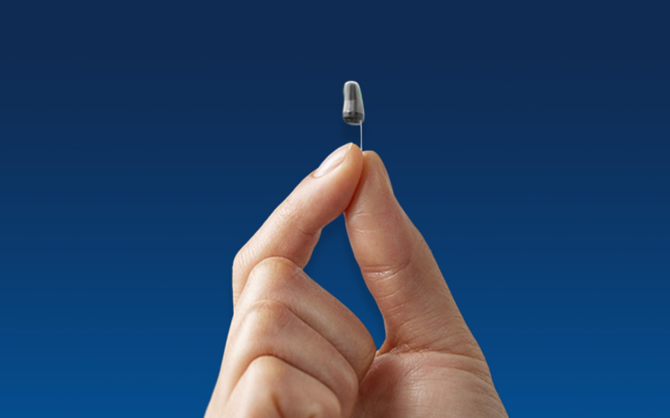 Very small Oticon Own Hearing Aid being held by two fingers