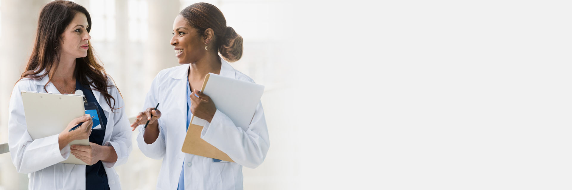 Two women in white laboratory coats are conversing and holding clipboards in a brightly lit, modern hallway. One gestures while speaking, the other listens attentively.