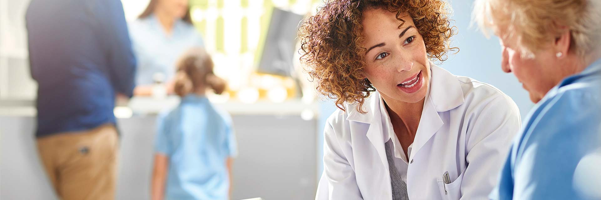 Doctor speaking with a patient in a clinic, with other patients and medical staff in the background engaging in various activities.