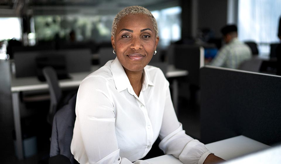 Person sitting and smiling at desk in an office, with blurred colleagues working at desks in the background.