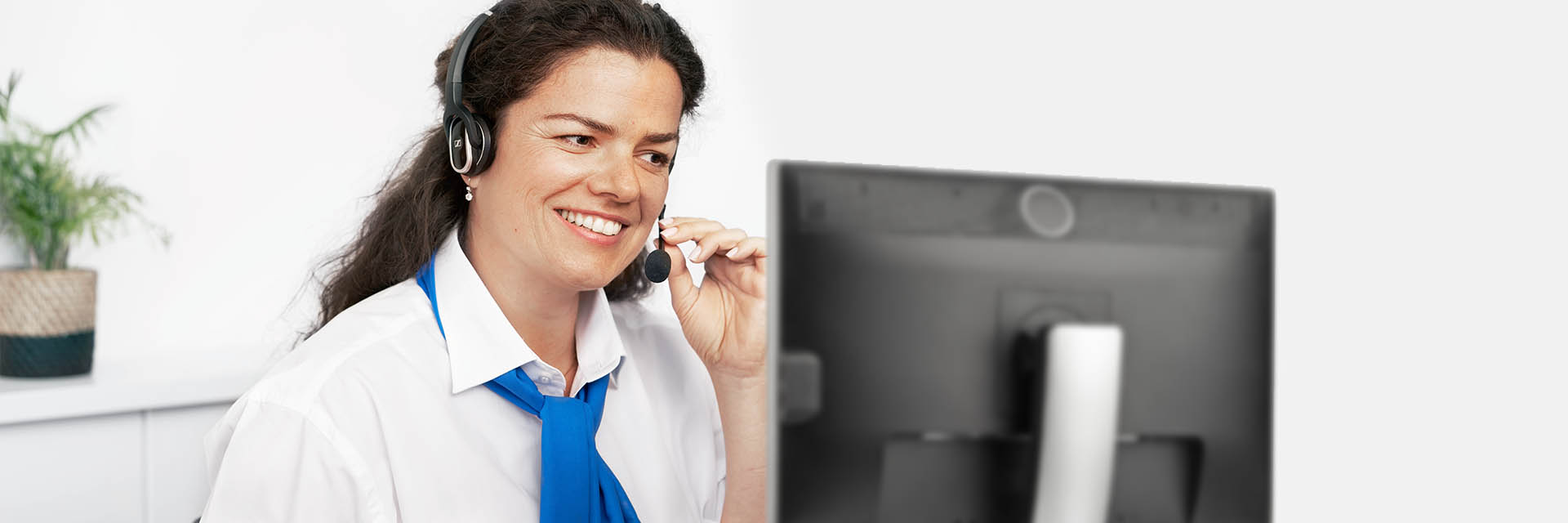 A person wearing a headset smiles while speaking into the microphone, seated in front of a computer monitor in a clean, well-lit office with a potted plant on a desk.