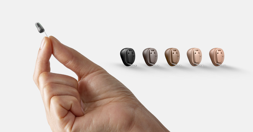 A hand holds a tiny hearing aid device, with five different colored hearing aids (black, brown, beige) displayed in a row on the right side against a white background.