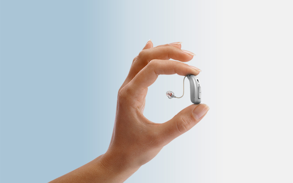 A hand holds a small hearing aid between the thumb and index finger; the background gradients from light blue to white.